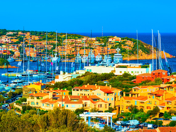 View of Porto Cervo marina in Sardinia, with luxury yachts docked along the waterfront, pastel-hued villas, and the iconic Piazzetta in the background.