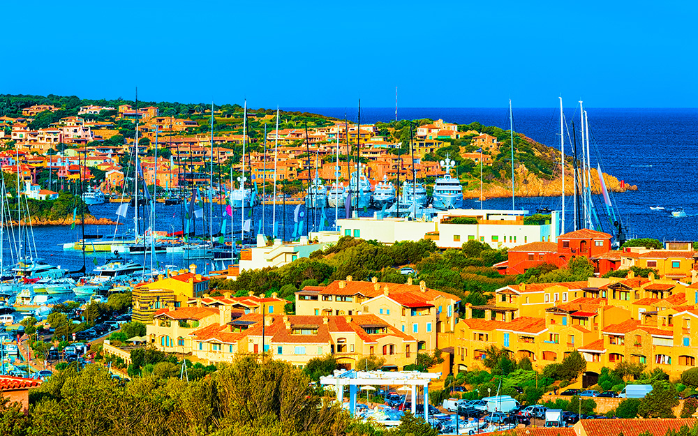 View of Porto Cervo marina in Sardinia, with luxury yachts docked along the waterfront, pastel-hued villas, and the iconic Piazzetta in the background.