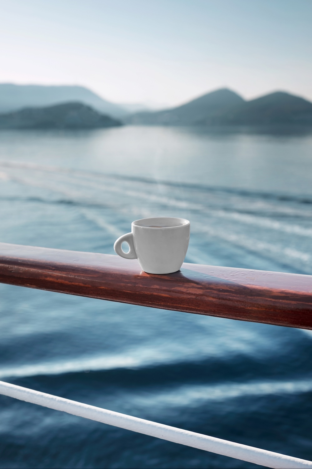White coffee cup resting on a yacht deck railing overlooking calm open sea and distant coastline