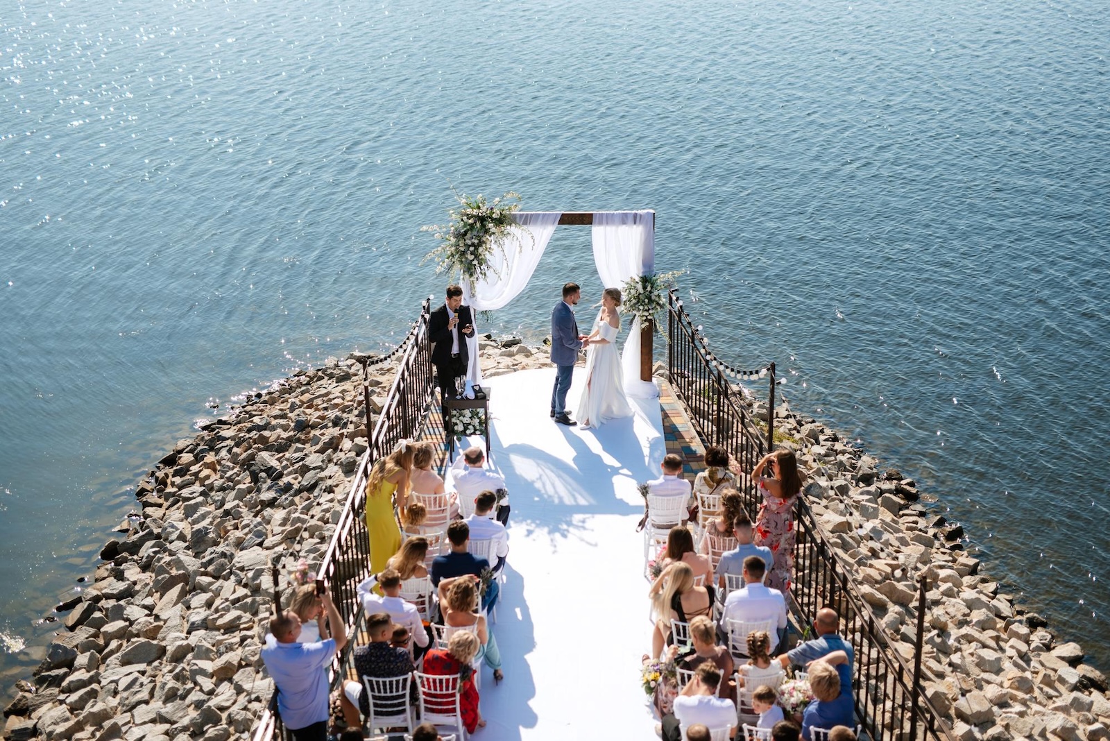 Intimate coastal wedding ceremony overlooking the sea with guests gathered beside the water