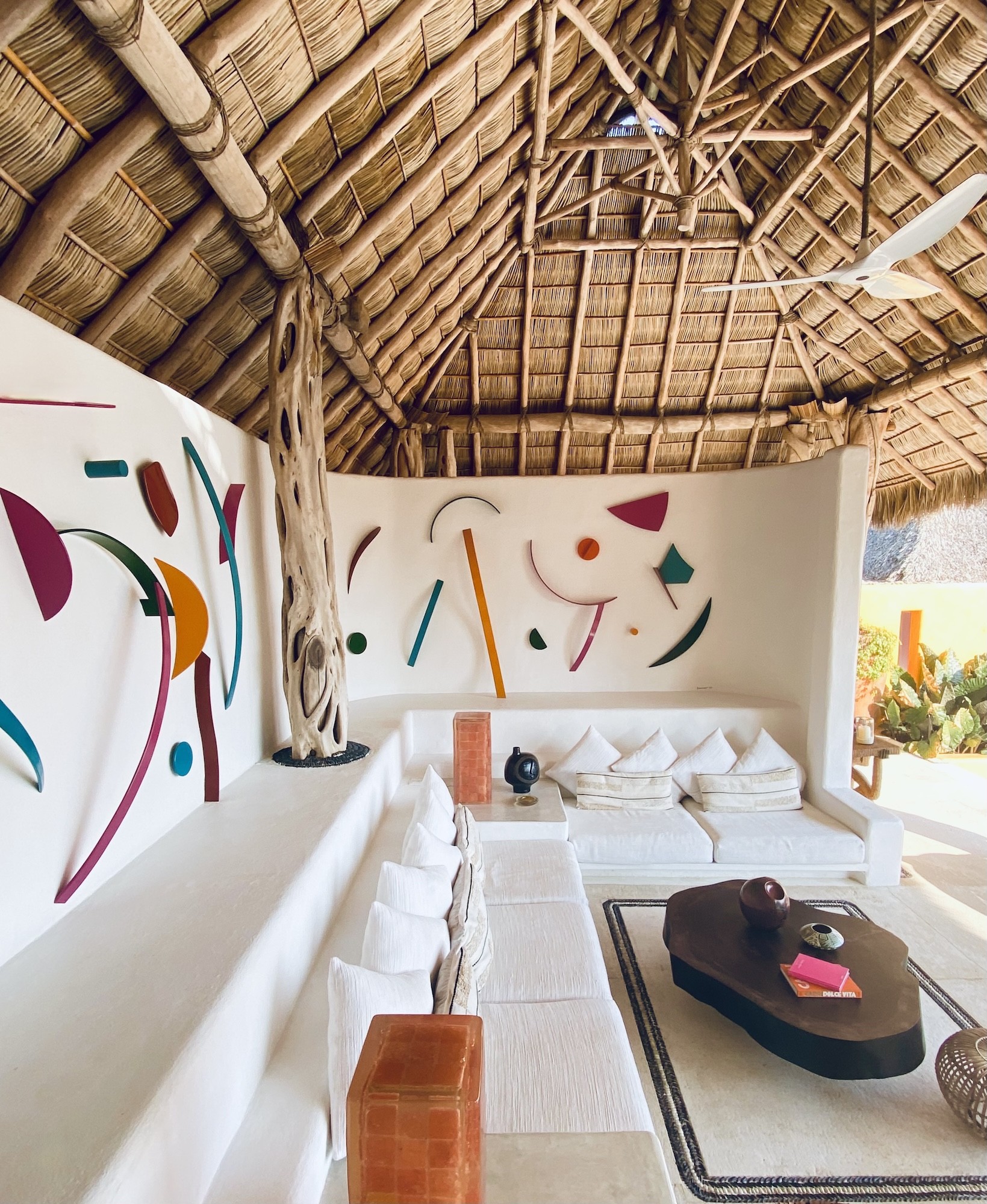 White living room with colourful abstract wall art beneath a natural palapa roof