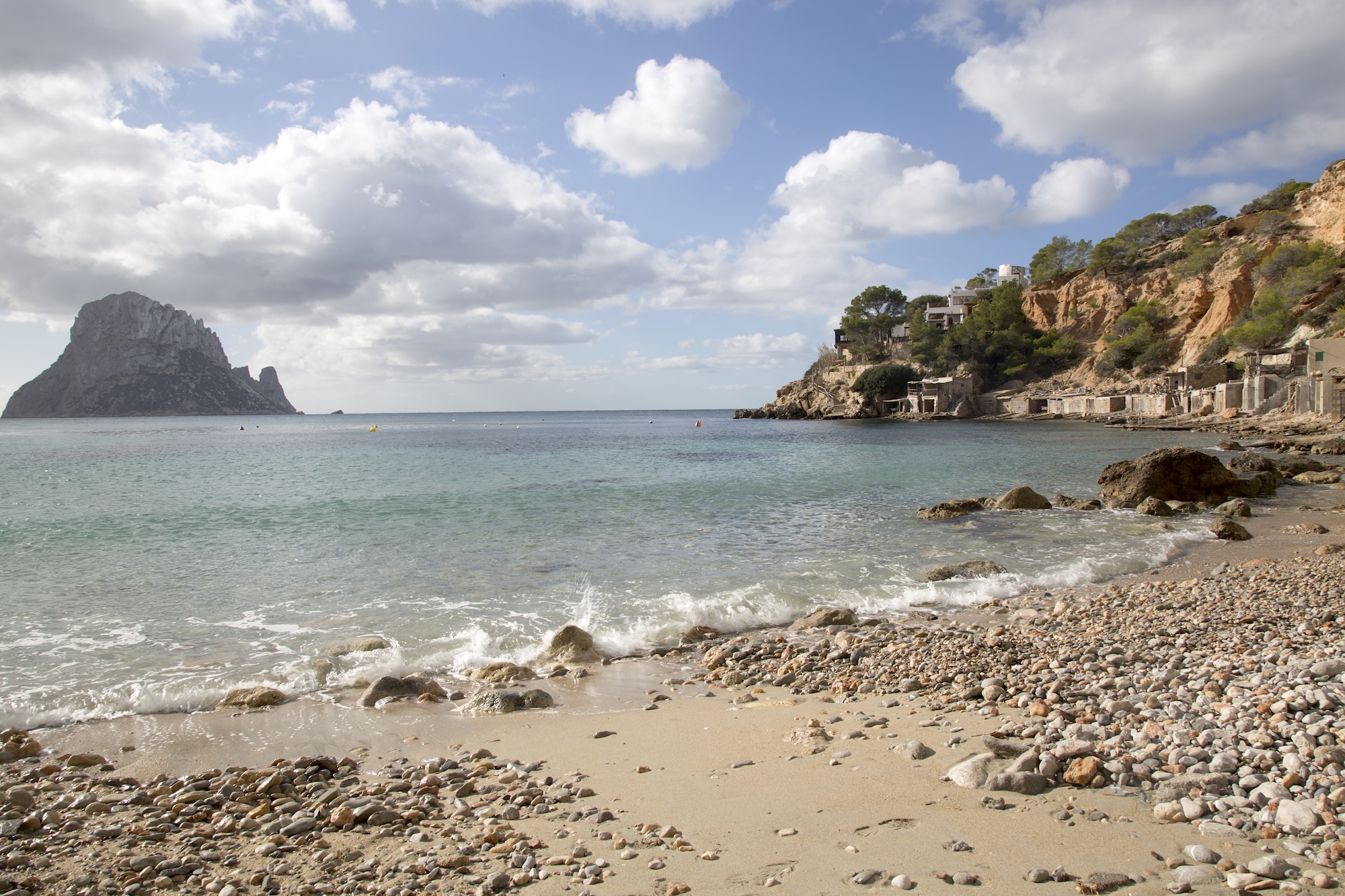 Sandy and pebbly shoreline at Cala d’Hort in Ibiza, with coastal cliffs and Es Vedrà offshore