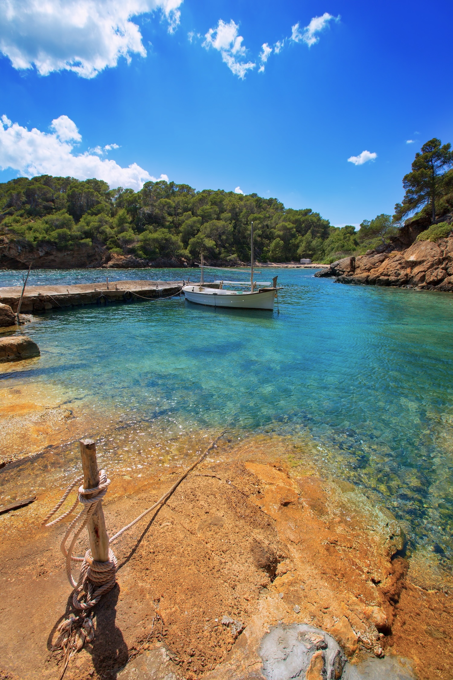 Small white boat moored in the clear turquoise water of Cala Mastella, Ibiza, beside a rocky, pine-lined shore