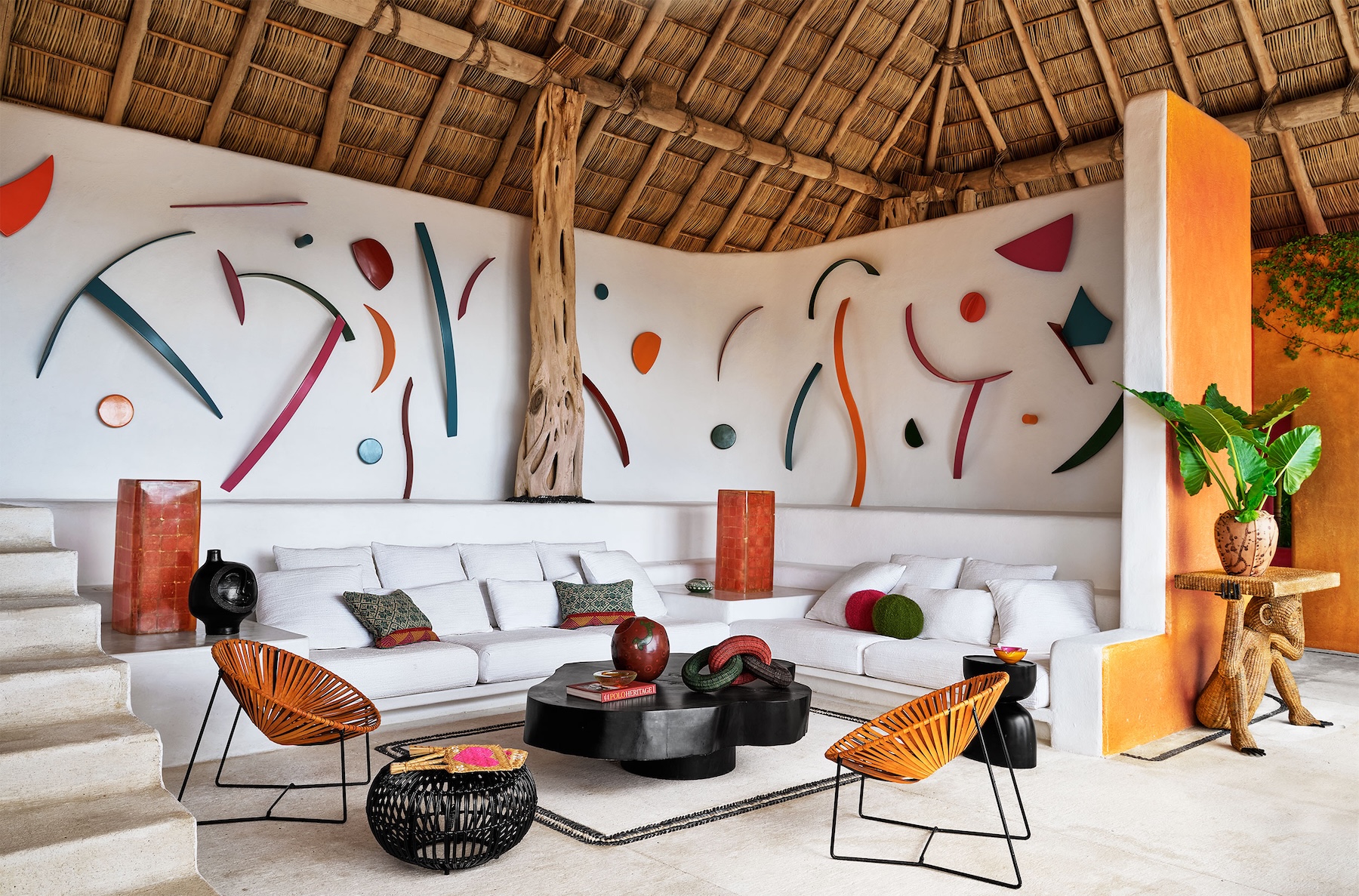Open-air palapa living room at Casa Torre with a thatched roof, white built-in seating, colourful wall art and woven chairs.
