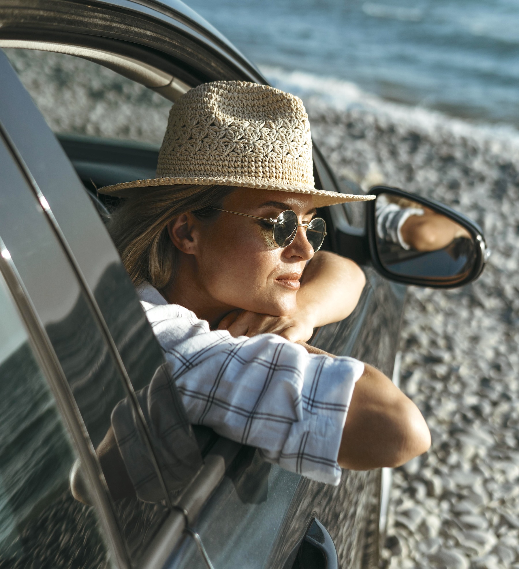 Woman wearing a straw hat and sunglasses leaning out of a car window beside the sea