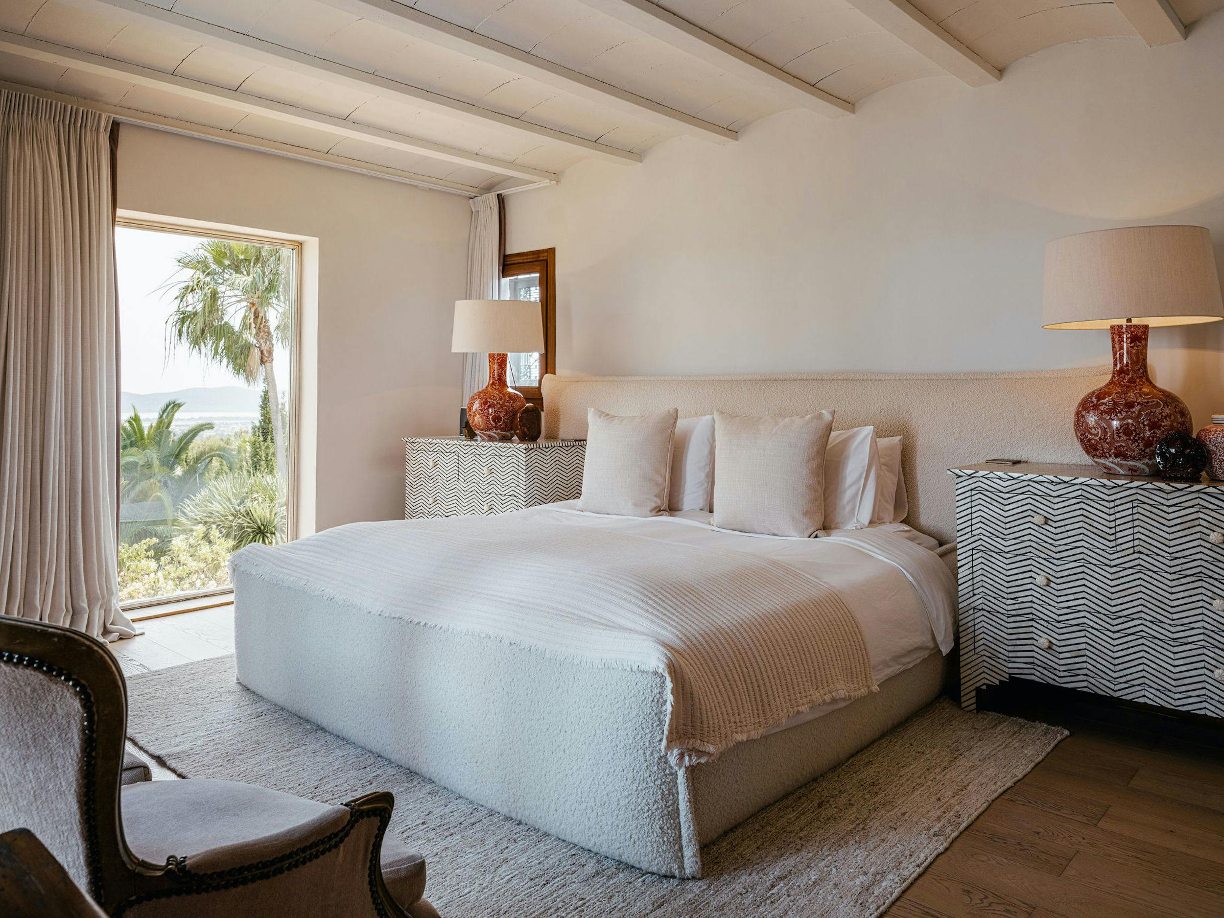 Bedroom at El Zafiro in Ibiza with a large upholstered bed, neutral furnishings, patterned bedside cabinets, and a window overlooking the gardens and sea.