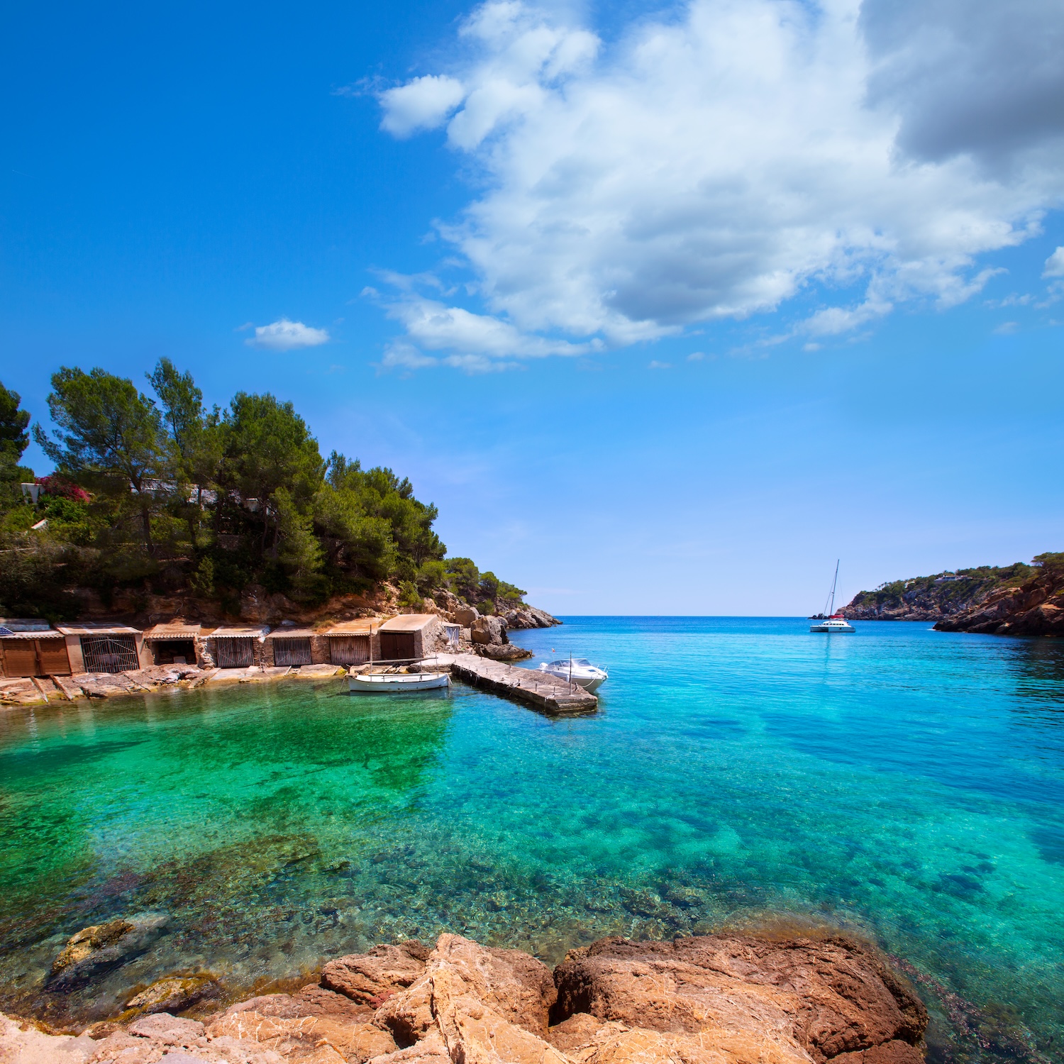 Cala Mastella in Ibiza with clear Mediterranean water, traditional boathouses and a sailing yacht