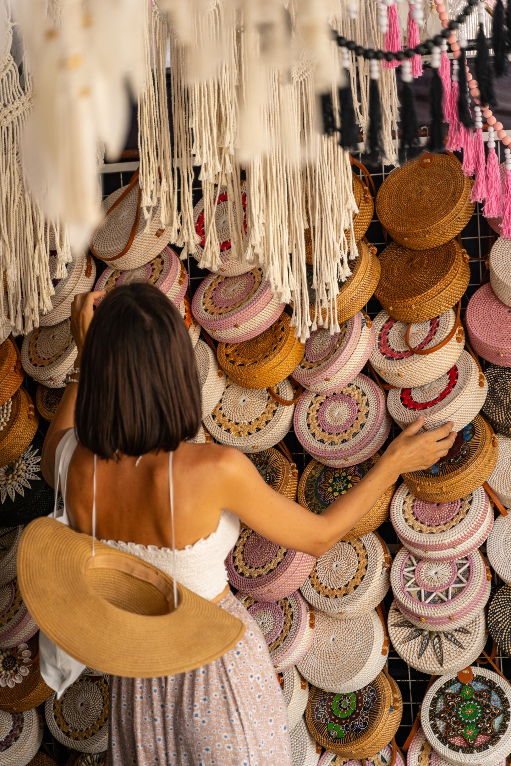 Woman browsing a display of woven bags and hanging textiles in a colorful Mediterranean-style shop.