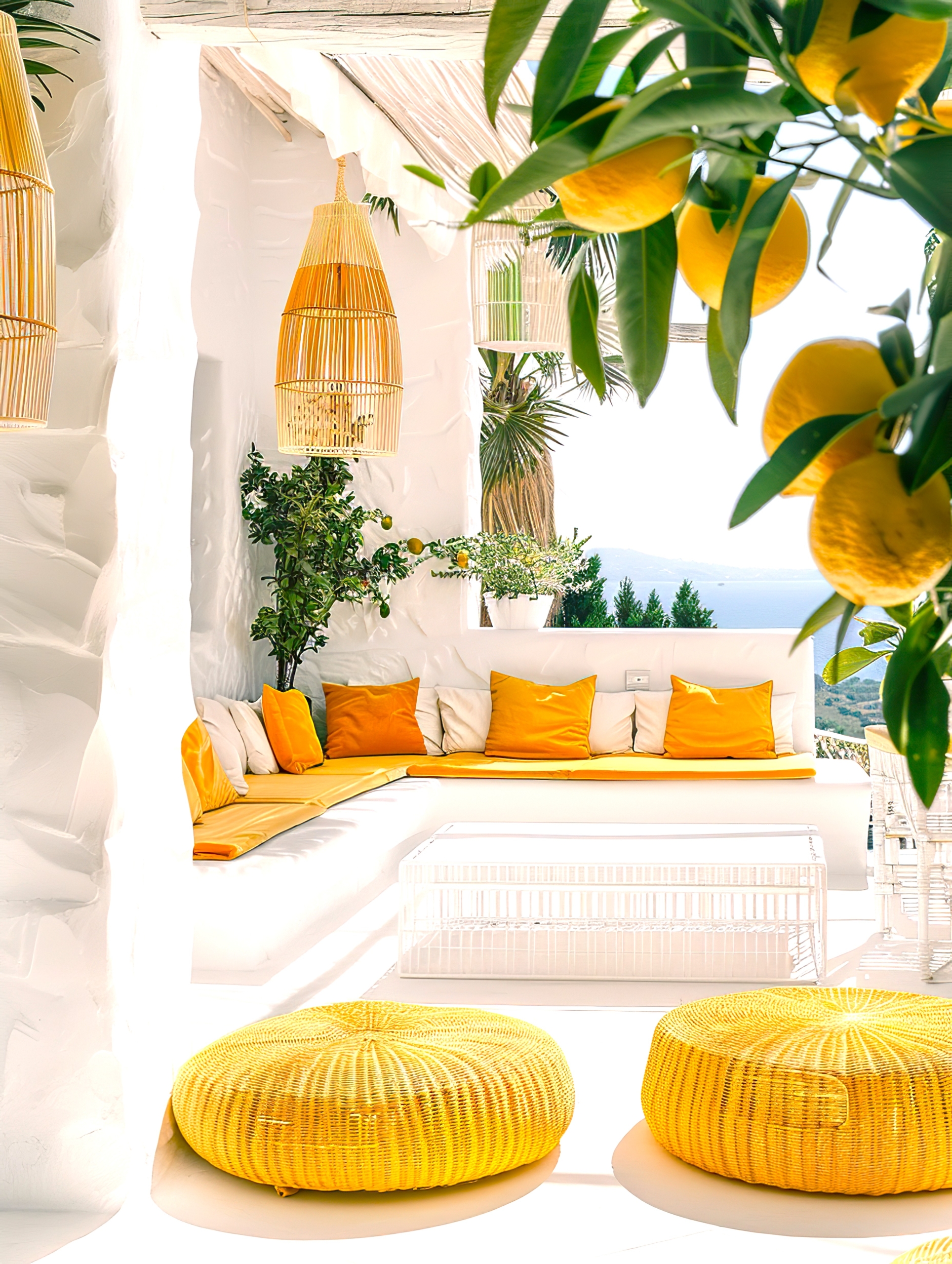 White Mediterranean terrace with cushioned seating, yellow accents, woven pendant lights, citrus foliage, and a view toward the sea