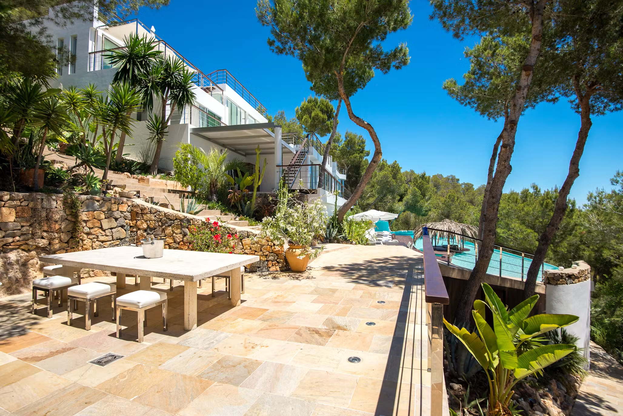Stone-paved outdoor dining terrace at Villa Destiny Ibiza, with a large dining table, pine trees and the swimming pool beyond.