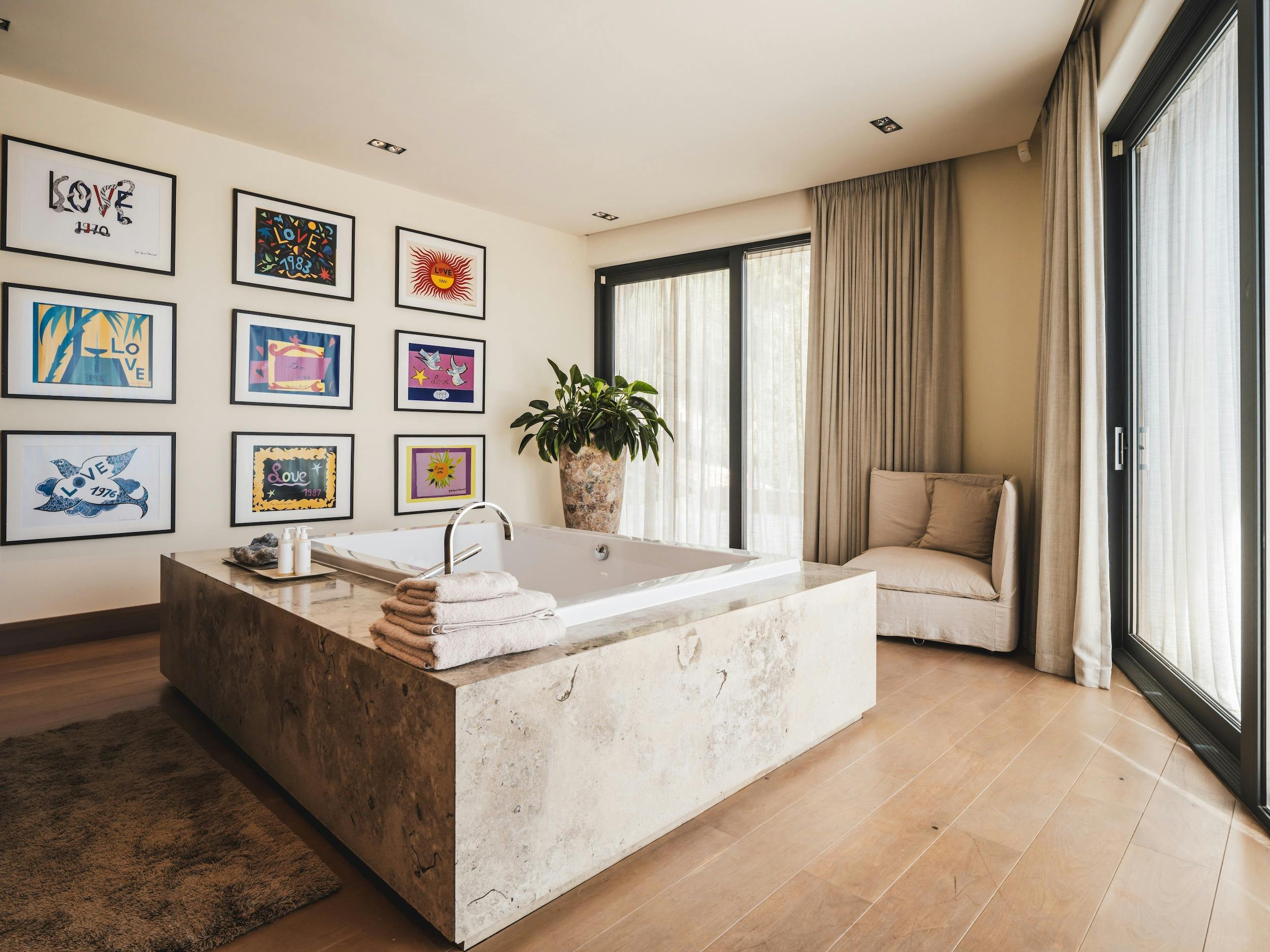 Large bathtub with a pale stone surround beside full-height curtained windows and colorful artwork at Ocean Paradise in Ibiza