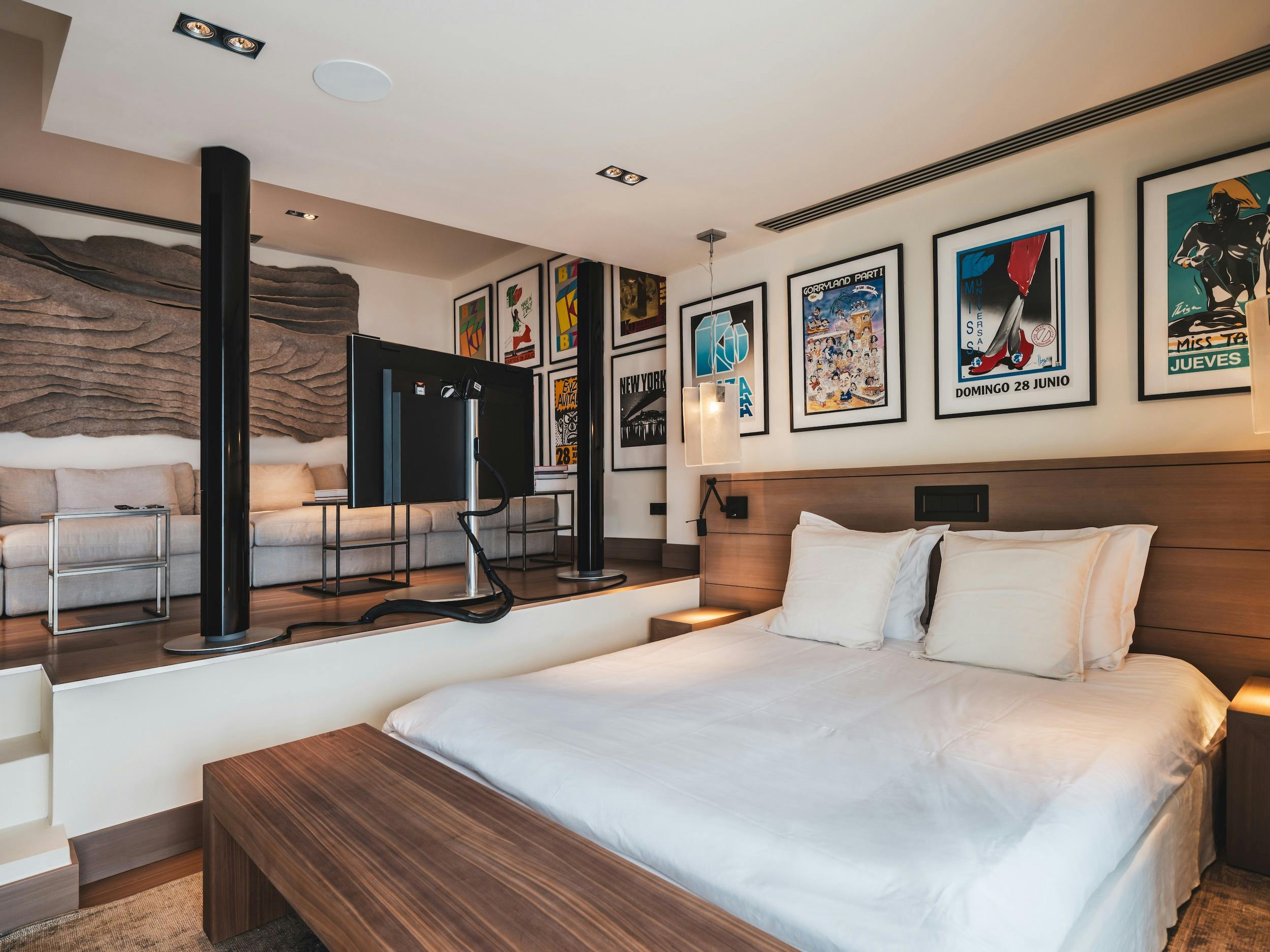 Bedroom with white bedding, framed posters, and a raised sitting area with a sofa and television at Ocean Paradise in Ibiza