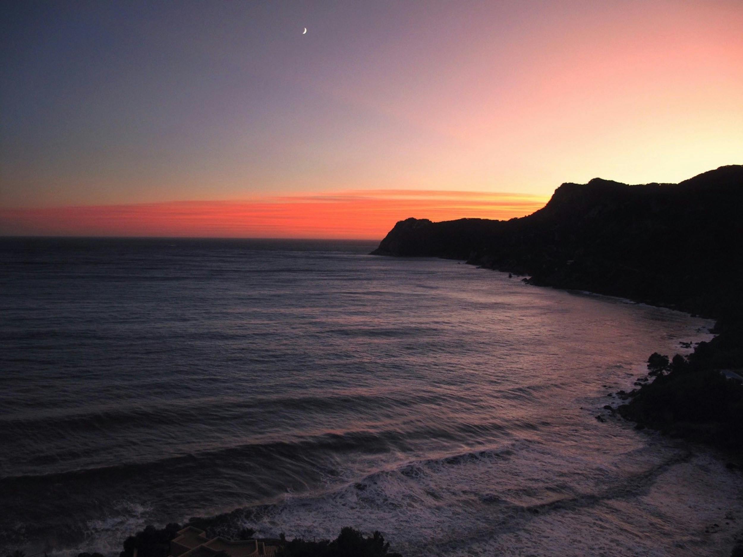 Pink and orange evening sky over the sea and silhouetted Ibiza coastline, with a crescent moon above