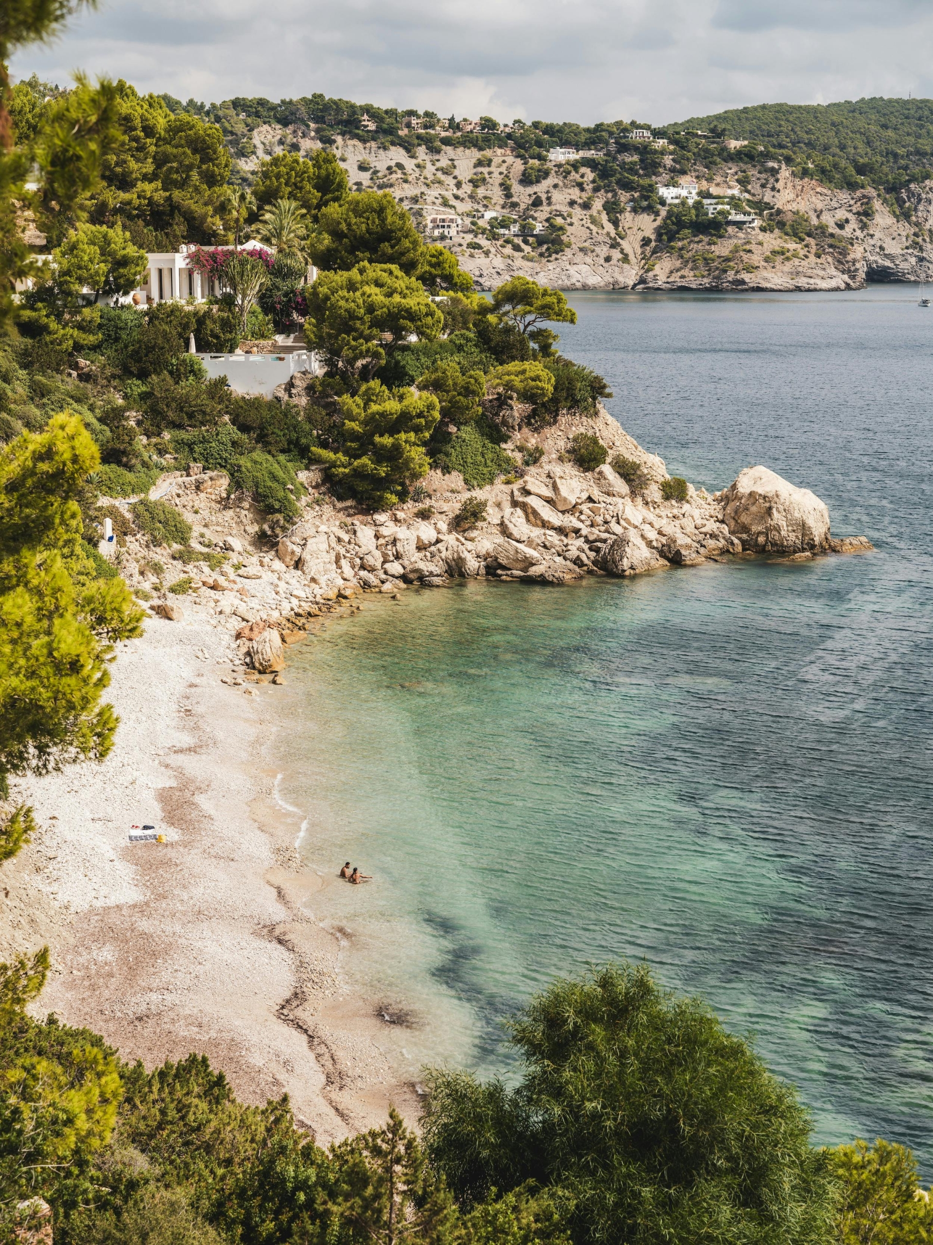 Pebble beach and clear turquoise water beneath pine-covered cliffs near Ocean Paradise Ibiza