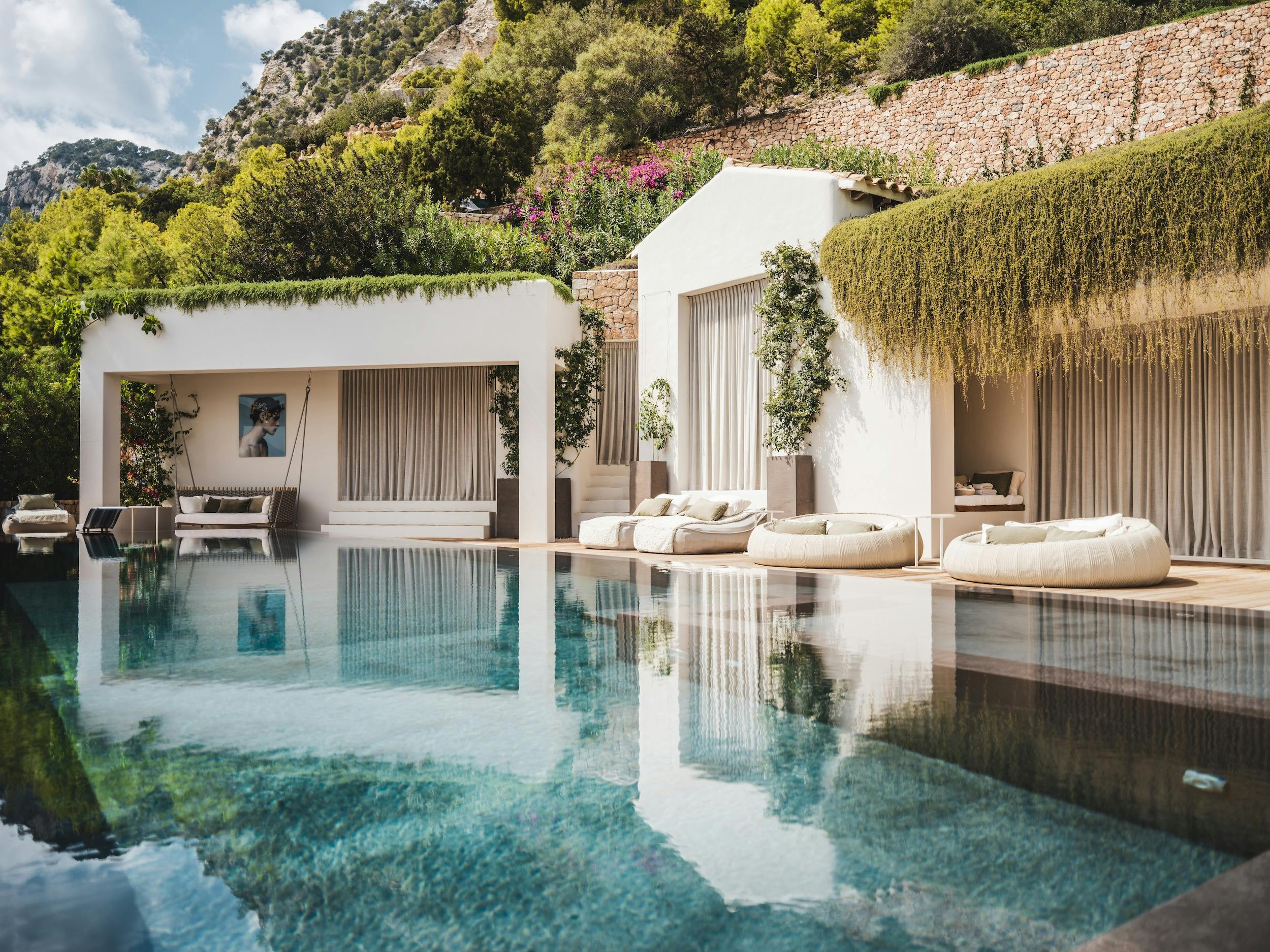 Poolside daybeds and a shaded swing beside the white villa at Ocean Paradise in Ibiza, backed by a green hillside