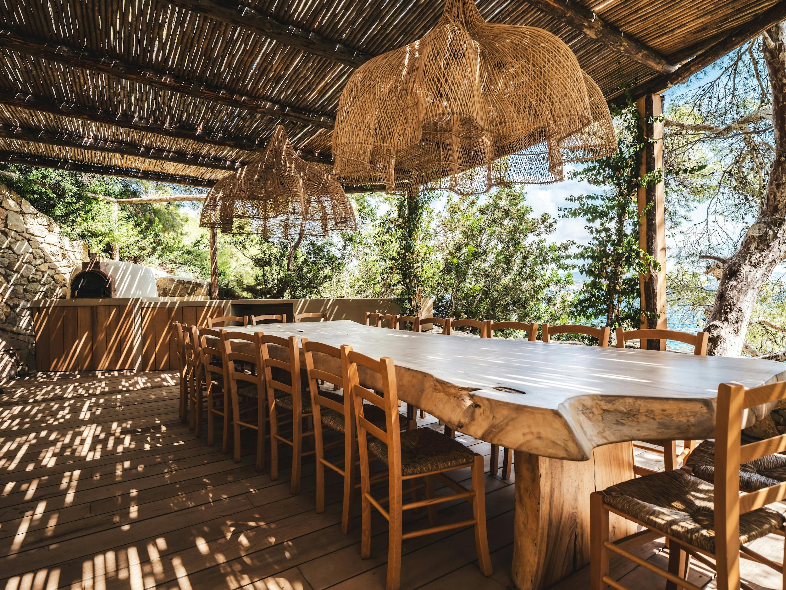 Long wooden dining table beneath a shaded pergola, with woven pendant lights and a pizza oven at Ocean Paradise in Ibiza
