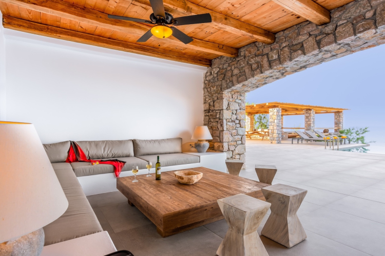 Shaded living room with timber ceiling and stone walls opening onto a sea-facing terrace in Crete