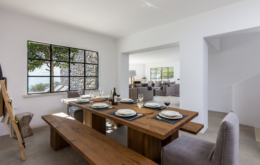 Large timber dining table in a white Mediterranean interior with black-framed window and sea view